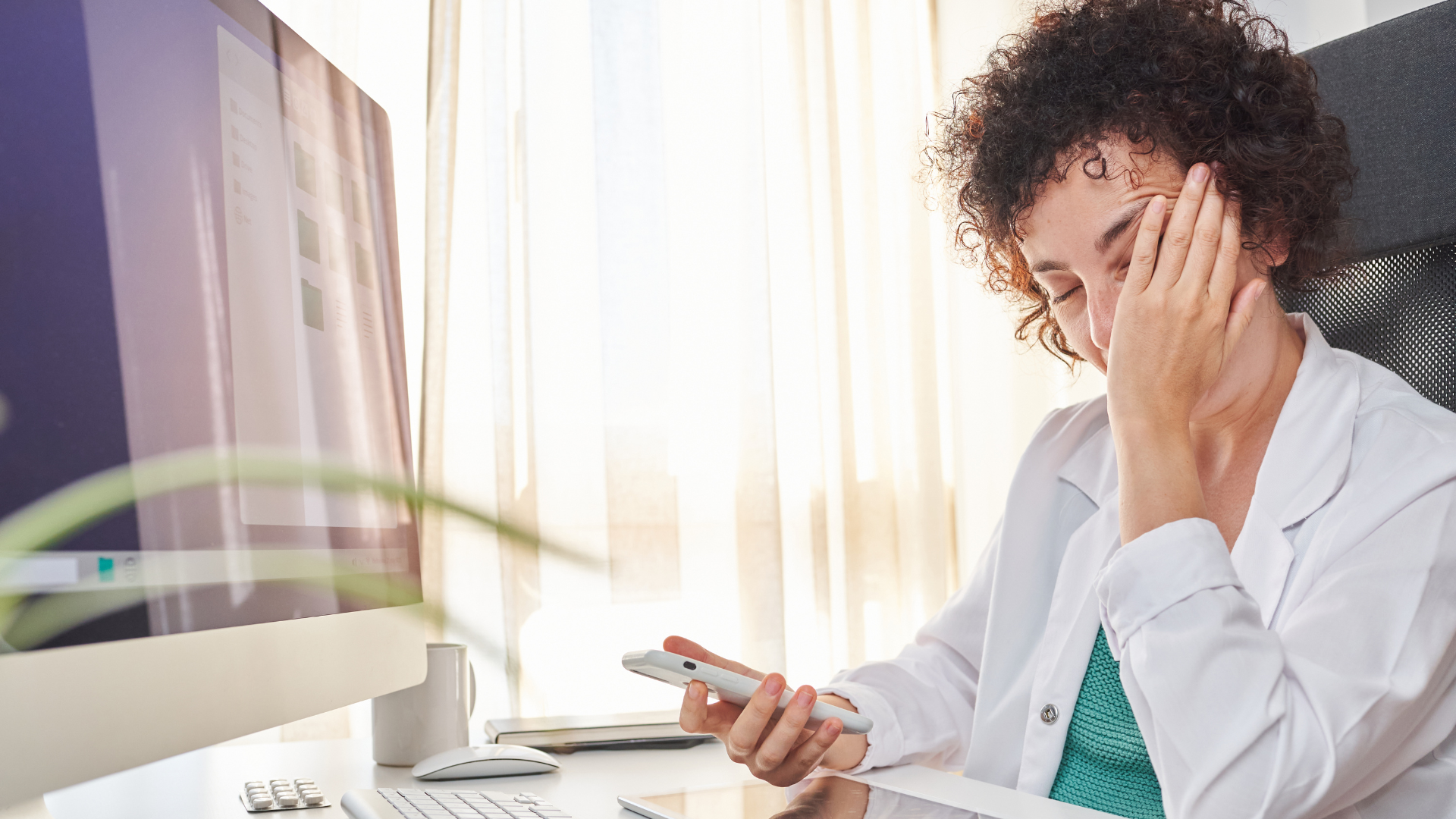 Burnout Behind the Scenes Exhausted doctor holding a phone, sitting at her desk with eyes closed in frustration.